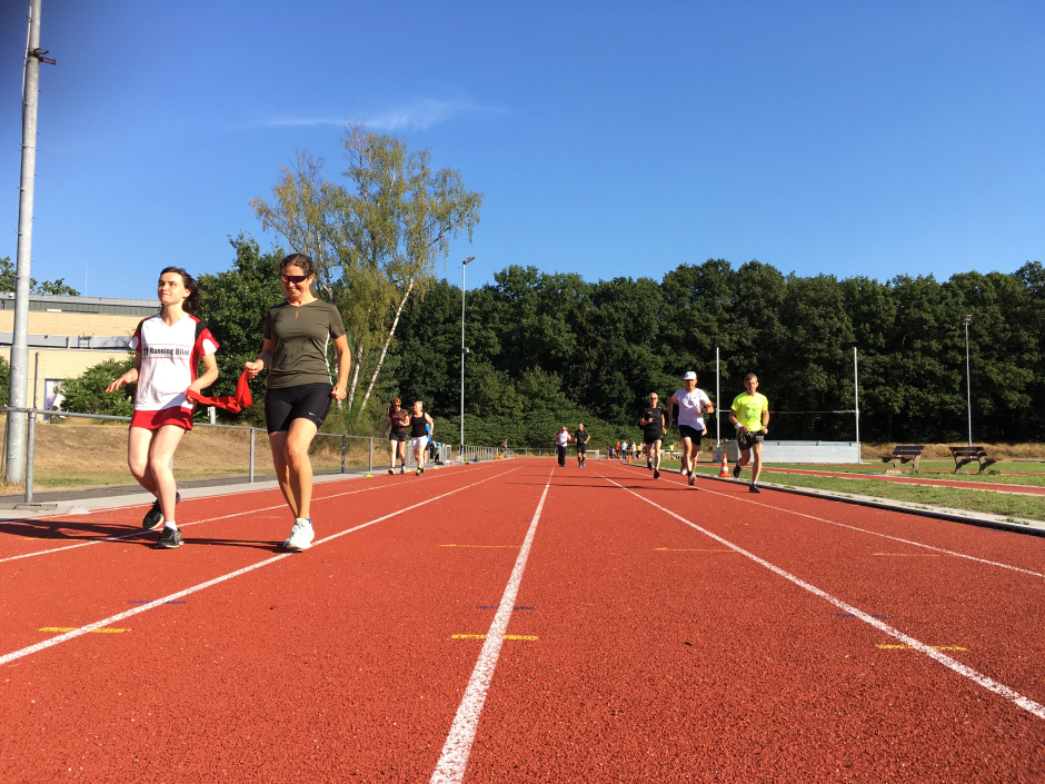 Running Blind-atleet en haar buddy lopen richting finish over atletiekbaan Dijnselburg. Op de achtergrond het clubhuis van Fit en de grote berkenboom voor een stralend blauwe hemel. 